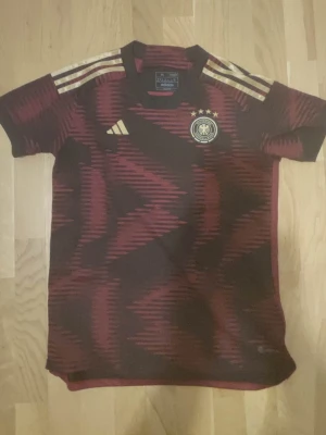 Tysklands matchtröja Adidas vinröd M - Säljer en vinröd Tyskland matchtröja från Adidas med guldiga detaljer och tre stjärnor ovanför landslagsemblemet. Tröjan har korta ärmar, rund hals och ett grafiskt mönster i mörkrött. Tillverkad i lätt och funktionellt material, perfekt för fotboll eller träning.