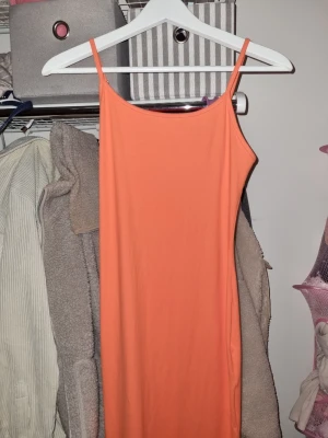 Persikofärgad lång klänning - En enkel och snygg persikofärgad lång klänning med smala axelband och rak passform. Klänningen är tillverkad i ett mjukt och stretchigt material som sitter skönt på kroppen. Perfekt för dig som gillar minimalistisk stil och vill ha något lätt att bära.