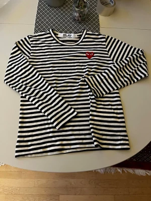 Randig långärmad tröja Comme des Garçons - Säljer en svartvit randig långärmad tröja från Comme des Garçons PLAY med det ikoniska röda hjärtat på bröstet. Tröjan har rund halsringning och är gjord i mjuk bomull. Perfekt för en stilren och trendig look.