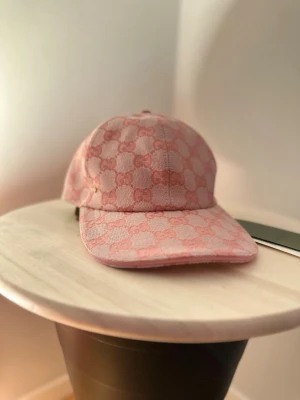 Rosa keps från Gucci med GG-mönster - Säljer en rosa keps Gucci med klassiskt GG-mönster över hela kepsen. Kepsen har böjd skärm och är tillverkad i ett tygmaterial som känns lyxigt och trendigt. Perfekt för dig som vill sticka ut med en designeraccessoar.
