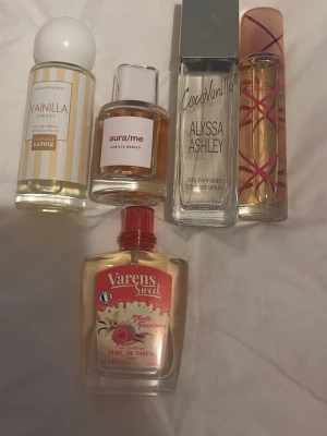 Fem olika vaniljparfymer mix - Fem flaskor parfym med vaniljdoft från olika märken: Saphir Vainilla Sweet, aura/me Vanilla Addict, Alyssa Ashley CocoVanilla, Varens Sweet Vanille Framboise och en okänd med rosa mönster. Flaskorna är i glas och plast, i olika former och färger, mestadels transparenta med vita, silver och rosa detaljer.
