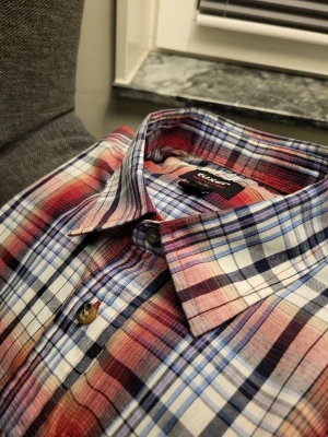 Rutig kortärmad skjorta från Tuxer - Kortärmad skjorta från Tuxer i rött, blått och vitt rutigt mönster. Skjortan har två bröstfickor med dragkedja och knappar framtill. Perfekt för dig som gillar en avslappnad och färgstark stil.