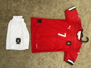 Portugal fotbollskit Ronaldo #7 - Snyggt Portugal fotbollskit med röd tröja och vita shorts. Tröjan har nummer 7 och namnet Ronaldo på ryggen, samt små mönster och gröna detaljer på ärmarna. Puma-logga på bröstet och shorts. Perfekt för fotbollsträning eller match.