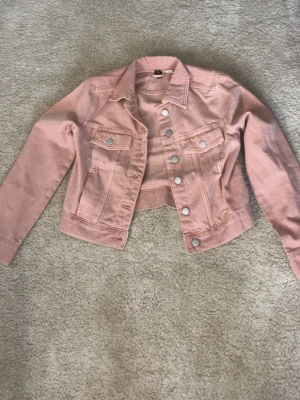 Ljusrosa cropped jeansjacka - Trendig ljusrosa jeansjacka med cropped passform och silvriga knappar framtill. Klassisk krage, två bröstfickor med lock och långa ärmar. Perfekt för att lyfta din outfit med en färgklick.