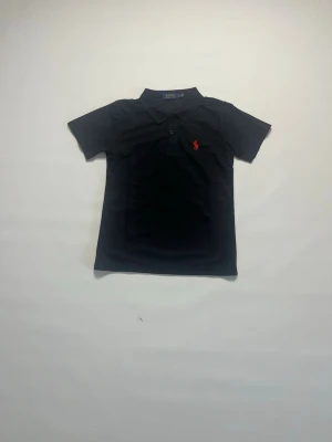 Svart pikétröja från Polo Ralph Lauren storlek S - Klassisk svart pikétröja från Polo Ralph Lauren med korta ärmar och röd broderad logga på bröstet. Tröjan har krage och knappar framtill, perfekt för en stilren look. Tröjan är storlek S och är helt ny och aldrig buren