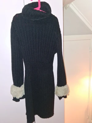 Svart stickad polotröja med fuskpäls - Mysig svart stickad polotröja med ribbad struktur och långa ärmar. Ärmsluten har breda, fluffiga detaljer i vit fuskpäls som ger en unik touch. Perfekt för kalla dagar när du vill vara både varm och snygg.