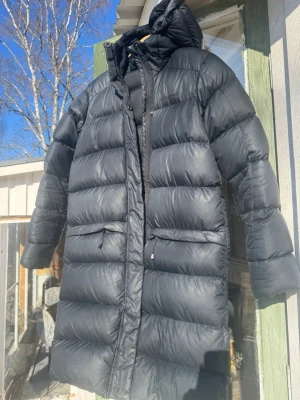 Svart lång dunjacka med huva - Snygg svart lång dunjacka med huva och dragkedja framtill. Jackan har två stora fickor med dragkedja och breda, quiltade paneler som ger en puffig look. Perfekt för kalla vinterdagar och riktigt varm.