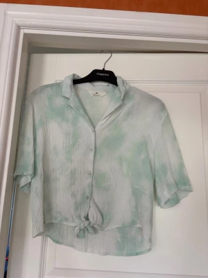 Grönvit knytblus från H&M - En luftig grönvit blus från H&M, storlek 158. Den har krage och korta ärmar och knytning och knappar framtill. Den har använts och det syns lite tecken på det, se sista bilden.