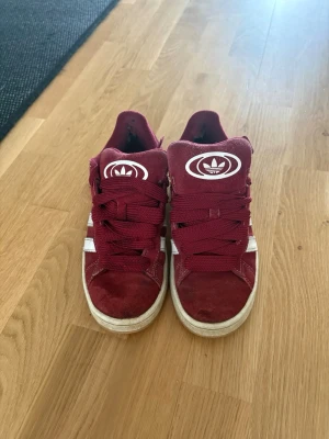 Vinröda Adidas sneakers i mocka - Snygga vinröda sneakers från Adidas med klassiska vita ränder på sidorna och stort Adidas-logga på plösen. Skorna har snörning och är gjorda i mocka för en cool och trendig look.