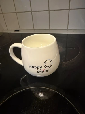 Vit mugg med smiley och text - Säljer en vit keramikmugg med rund form och stort handtag. Ena sidan har texten 'Happy smile!!' och en liten smiley, andra sidan har ett stort leende ansikte. Perfekt för te eller kaffe och ger en glad vibe till frukosten.