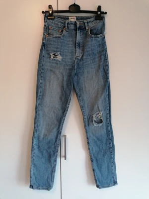 High waist slim leg jeans från ASOS - Ett par ljusblå high waist slim legs jeans från ASOS. Jeansen har slitna detaljer på ena låret och knät samt klassiska fem fickor. Perfekta för en avslappnad och trendig look.