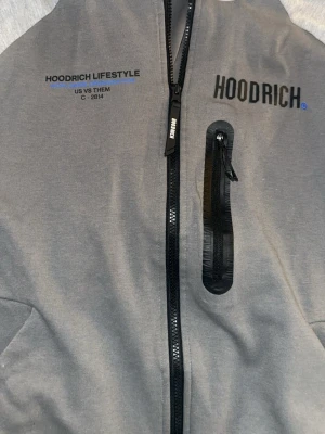 Grå hoodie från Hoodrich med dragkedja - Snygg grå hoodie från Hoodrich med svart dragkedja framtill och en extra bröstficka med dragkedja. Tryck på bröstet med texten 'HOODRICH LIFESTYLE' och 'HOODRICH'. Perfekt för en streetwear-look.