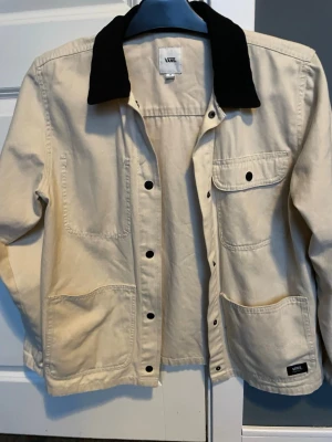 Beige overshirt från Vans med svart krage - Stilren beige overshirt från Vans med kontrasterande svart krage. Jackan har tryckknappar framtill, tre stora fickor och en bröstficka med knapp. Tillverkad i kraftigt bomullstyg som ger en avslappnad men snygg look.