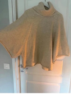 Beige poncho med polokrage - Snygg beige poncho med hög polokrage och vida ärmar. Ponchon är stickad i mjukt material. Väldigt trendig från Zara. Inga fläckar eller defekter storlek S