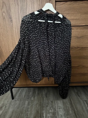 Svart prickig blus från Gina Tricot - Snygg svart blus med vita prickar från Gina Tricot. Blusen har långärmade, luftiga ärmar och en knytning i halsen. Tillverkad i ett lätt, transparent tyg som ger en cool och trendig look. Perfekt att styla med jeans eller kjol.