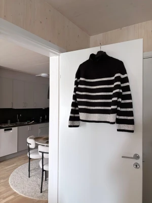 Randig svartvit stickad polotröja - Snygg stickad polotröja med breda svarta och vita ränder. Tröjan har hög krage och långa ärmar, perfekt för kyliga dagar. Den är ribbstickad i muddar och nederkant för extra komfort och stil.