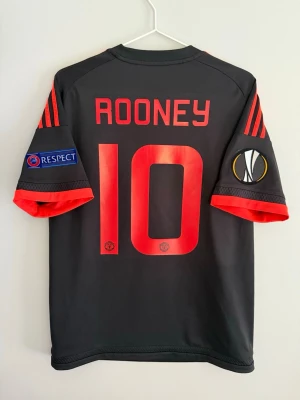 Manchester United Rooney matchtröja - Säljer en svart Manchester United matchtröja från Adidas med röda detaljer, nummer 10 och Rooney på ryggen. Tröjan har officiella Europa League och Respect-patchar på ärmarna samt klubbmärke och sponsorlogga framtill. Storlek: M Skick: 8/10 Produktkod: AC1445