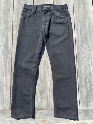 Nudie Jeans Hank Rey - Org. Dry Black - Säljer ett par Nudie Jeans i modellen Hank Rey. Modellen tillverkas inte längre. Jeansen är i färgen "Org. Dry Black" och är gjorda i 100% ekologisk bomull. De är i mycket fint skick med en djup, matt svart ton.  Modell: Hank Rey Färg: Org. Dry Black Storlek: W31 / L32  Exakta mått: Jag har mätt jeansen plant för att säkerställa att de passar dig:  Midja: 40 cm  Innerbenslängd (från grenen): 70 cm  Ytterbenslängd: 99 cm  Lår: 25 cm  Benöppning: 17 cm  Skickas inom 1-2 dagar