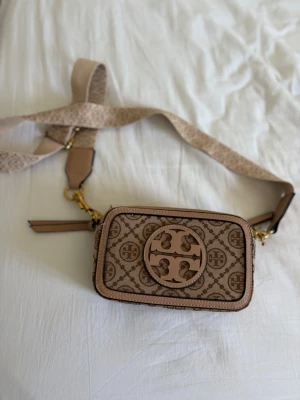 Beige axelremsväska från Tory Burch ej äkta - Snygg beige handväska från Tory Burch med justerbar axelrem och stort logomönster framtill. Väskan är rektangulär och har ett mönstrat tygmaterial med läderdetaljer och guldfärgade metalldelar. Perfekt för dig som gillar stilrena accessoarer. Ej äkta