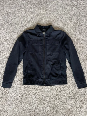 Svart overshirt från Jack & Jones - Stilren svart overshirt från Jack & Jones med dragkedja framtill, två bröstfickor och klassisk krage. Materialet är mjukt och lätt, perfekt för lager-på-lager. Passar dig som gillar en clean och enkel look.