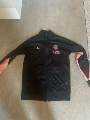 PSG x Jordan hoddie  - Svart träningsjacka från PSG x Jordan med rosa detaljer på ärmarna och loggor på bröstet. Jackan har dragkedja framtill, två fickor med dragkedja och är tillverkad i Dri-FIT-material som andas. Perfekt för fotbollsträning eller chill.