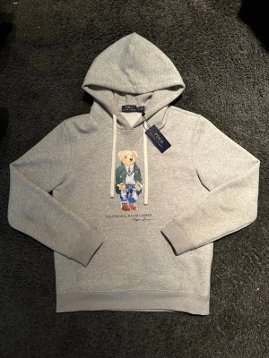 Grå hoodie Polo Bear Ralph Lauren - Storlek M passar S också .  .  Snygg grå hoodie från Ralph Lauren med den ikoniska Polo Bear-trycket framtill. Hoodien har dragsko i huvan och ribbade muddar vid ärmar och nederkant. Perfekt för dig som gillar streetwear och klassisk stil. Materialet känns mjukt och bekvämt. Obs prislapp följer ej med. 