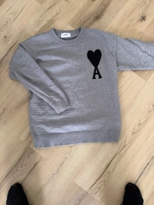 Grå tröja med svart hjärta från AMI - Snygg grå tröja från AMI med ett stort svart hjärta och bokstaven A broderat på bröstet. Tröjan har rund hals, ribbade muddar och är tillverkad i mjukt material som passar perfekt för en chill look.