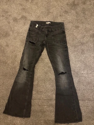 Svarta bootcut jeans med slitningar - Säljer ett par svarta bootcut jeans med slitningar över knäna och lätt tvättad look. Jeansen har två knappar i midjan och framfickor. Perfekta för en trendig och avslappnad stil.