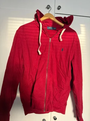 Röd hoodie från Polo Ralph Lauren - Röd hoodie med dragkedja från Polo Ralph Lauren. Klassisk design med huva och vita dragsnören, två fickor framtill och broderad logga på bröstet. Perfekt för en avslappnad och stilren look.