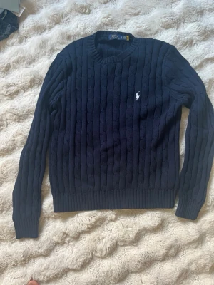 Mörkblå kabelstickad tröja Polo Ralph Lauren - Mörkblå kabelstickad tröja från Polo Ralph Lauren med rund halsringning och lång ärm. Tröjan har knappt använts och ser helt sprillans ny ut utan några skador eller hål.