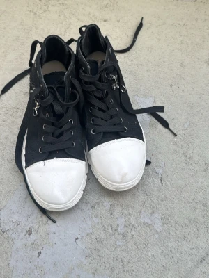 Svarta high top sneakers med hjärtberlocker - Säljer ett par svarta high top sneakers med vita tåpartier och tjock vit sula. Skorna har svarta snören, metallöljetter och detaljer som små hjärtformade berlocker vid dragkedjan på sidan. Materialet är canvas och sulan är i gummi.