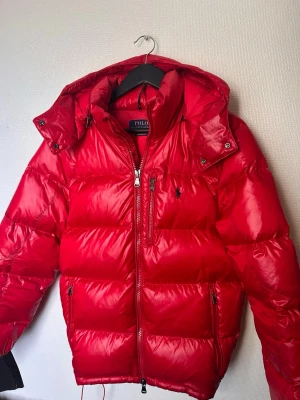 Röd pufferjacka från Polo Ralph Lauren - Snygg röd pufferjacka från Polo Ralph Lauren med glansig finish, avtagbar huva och dragkedjor framtill. Jackan har flera fickor med dragkedja och ikonisk broderad logga på bröstet. Perfekt för kalla vinterdagar.
