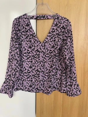 Lila blommig blus från Gina Tricot - Supersöt blus från Gina Tricot med lila blommönster på svart botten. Djup V-ringning och långa ärmar med lätt utsvängda ärmslut. Tunt och flowigt material som ger en snygg siluett. Perfekt att styla med jeans eller kjol.