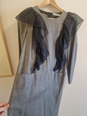 Grå klänning med volang från Rodebjer - Snygg grå klänning från Rodebjer med svarta volangdetaljer i transparent tyg framtill. Klänningen har lång ärm och rak passform, perfekt för dig som gillar stilrena plagg med en twist.