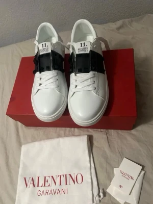 Valentino Rockstud Untitled sneakers - Snygga vita sneakers från Valentino Garavani med svarta detaljer och ikoniska Rockstuds på sidan. Skorna har klassisk låg profil, vita skosnören och är tillverkade i slätt läder. Perfekta för dig som vill ha en stilren men edgy look. BOX INGÅR TYVÄRR INTE, däremot ingår påsen!