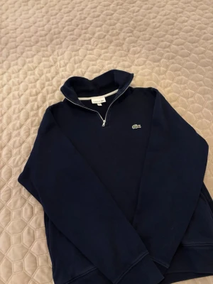 Mörkblå ziptröja från Lacoste - Snygg mörkblå ziptröja från Lacoste med halv dragkedja och klassisk krokodillogga på bröstet. Tröjan har hög krage och är tillverkad i mjukt bomullsmaterial. Perfekt för en stilren och avslappnad look.
