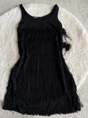 Svart fransklänning från Cubus - Supersnygg svart klänning från Cubus med fransdetaljer som ger en cool och festlig look. Klänningen är ärmlös och har en kroppsnära passform med rund halsringning. Perfekt för dig som vill sticka ut med en unik stil.