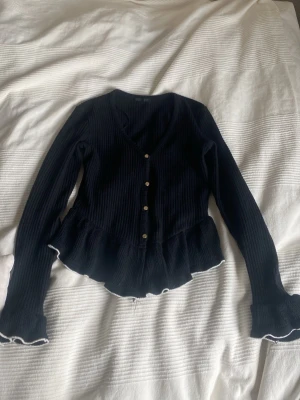 Svart ribbad kofta med volangkant - Snygg svart ribbad kofta med v-ringning och knappar framtill. Koftan har volangkant nedtill och vid ärmsluten, samt vita detaljer längs kanterna. Perfekt för dig som gillar en feminin och trendig stil.