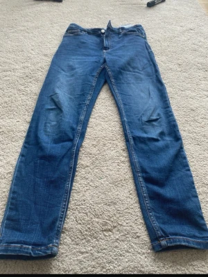 Blå raka jeans i klassisk modell - Säljer ett par blå jeans med rak passform och klassisk femficksdesign. Jeansen har en snygg tvätt med lätt slitning vid knäna och är tillverkade i denim. Perfekta för en avslappnad och stilren look.