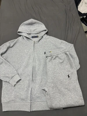 Grått set från Polo Ralph Lauren - Snyggt grått set från Polo Ralph Lauren med zip-hoodie och matchande mjukisbyxor. Båda plaggen har den klassiska broderade loggan i svart. Hoodien har dragkedja och fickor, byxorna har resår i midjan och sidofickor. Perfekt för en chill och avslappnad stil.
