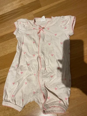 Rosa babyshortsdräkt från H&M - Supersöt pyjamas från H&M i vit färg med små rosa hjärtan. Kortärmad modell med tryckknappar framtill och en liten rosett vid halsen. Rosa dekorationssömmar längs kanterna. Endast använd på dockor, lite nopprig men inget som påverkar varan. Skickar helst med PostNord😃