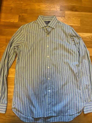 Randig blå skjorta från Polo Ralph Lauren - Snygg blå skjorta med vita och ljusgula ränder från Polo Ralph Lauren. Klassisk krage, långärmad och knäppning framtill. Perfekt för en stilren look och passar till många olika outfits.  Strl 15 1/2 35 ~ M