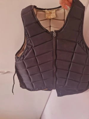 Svart skyddsväst med dragkedja - Svart vadderad skyddsväst med dragkedja framtill och snörning i sidorna. Västen har en sportig look och är fodrad i beige på insidan. Perfekt för aktiviteter där extra skydd behövs.