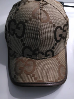 Beige GG-keps från - Säljer en beige keps med det klassiska GG-mönstret i brunt och svart. Kepsen har böjd skärm med brun kant och justerbart band bak i brunt läder med guldfärgade detaljer. Perfekt för dig som gillar exklusiva accessoarer och streetstyle.