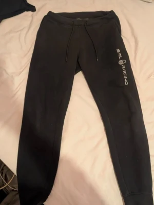 Svarta mjukisbyxor Sail Racing - Svarta mjukisbyxor från Sail Racing med dragsko i midjan och tryckt logga längs ena benet. Byxorna har smal passform, resår vid benslut och en bakficka. Perfekta för chill eller träning.