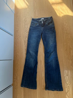 Mörkblå bootcut jeans med brodyr - Snygga mörkblå bootcut jeans med klassisk femficksdesign och broderade detaljer på bakfickorna. Jeansen har låg midja och utsvängda ben. Perfekta för dig som gillar en retrostil. Materialet är denim i bomull.