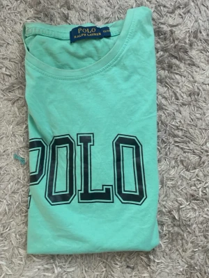 Mintgrön t-shirt från Polo Ralph Lauren - Fräsch mintgrön t-shirt från Polo Ralph Lauren med stor POLO-text i svart på bröstet. Klassisk rund halsringning och korta ärmar. Perfekt för dig som gillar stilrena och sportiga plagg med tydlig logga.