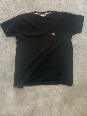 Svart t-shirt från Lacoste - Klassisk svart t-shirt från Lacoste med den ikoniska gröna krokodilloggan broderad på bröstet. T-shirten har rund halsringning och korta ärmar. Tillverkad i mjuk bomull för en bekväm känsla och enkel stil.
