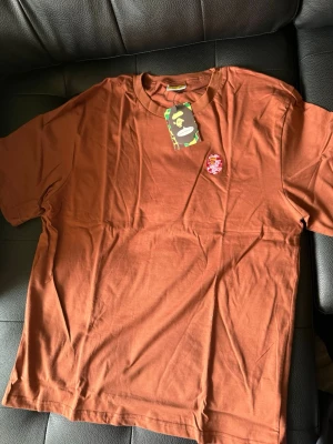 Brun t-shirt från A Bathing Ape - Säljer en brun t-shirt från A Bathing Ape med klassisk rund hals och korta ärmar. På bröstet finns en broderad logga i rött och vitt. Perfekt för dig som gillar streetwear och vill sticka ut lite extra.