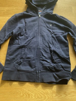 Mörkblå hoodie från Gant - Mörkblå hoodie från Gant med dragkedja framtill och två stora fickor. Klassisk huva och diskret Gant-logga broderad på bröstet. Tillverkad i mjuk bomullsblandning.